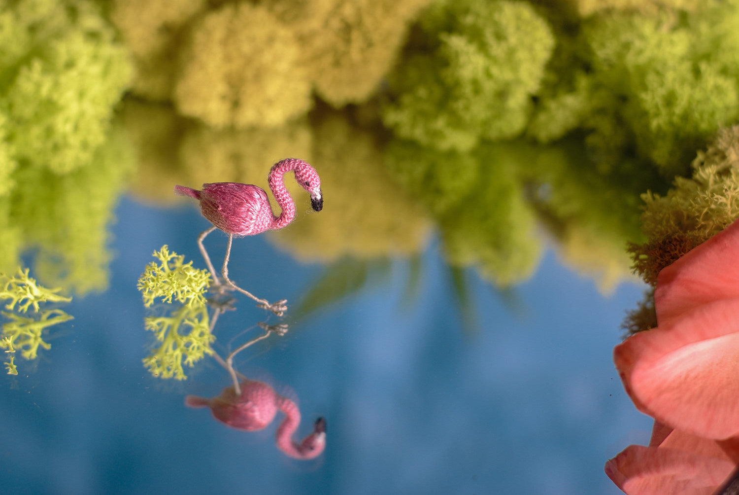 Miniature crochet flamingo reflected in water, handmade micro bird in a natural green setting