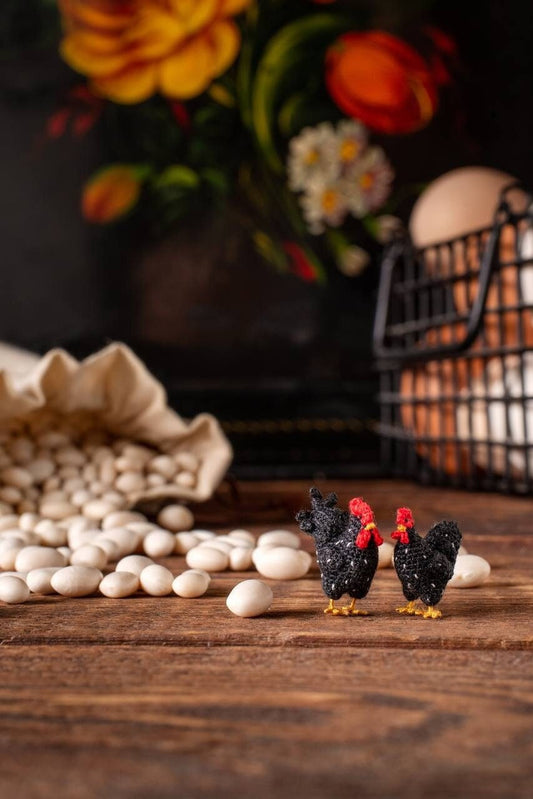 tiny handmade crochet chickens in black with red crests, miniature rooster and hen pair standing on rustic table with beans and eggs in background, TaleWorld Miniature Creatures
