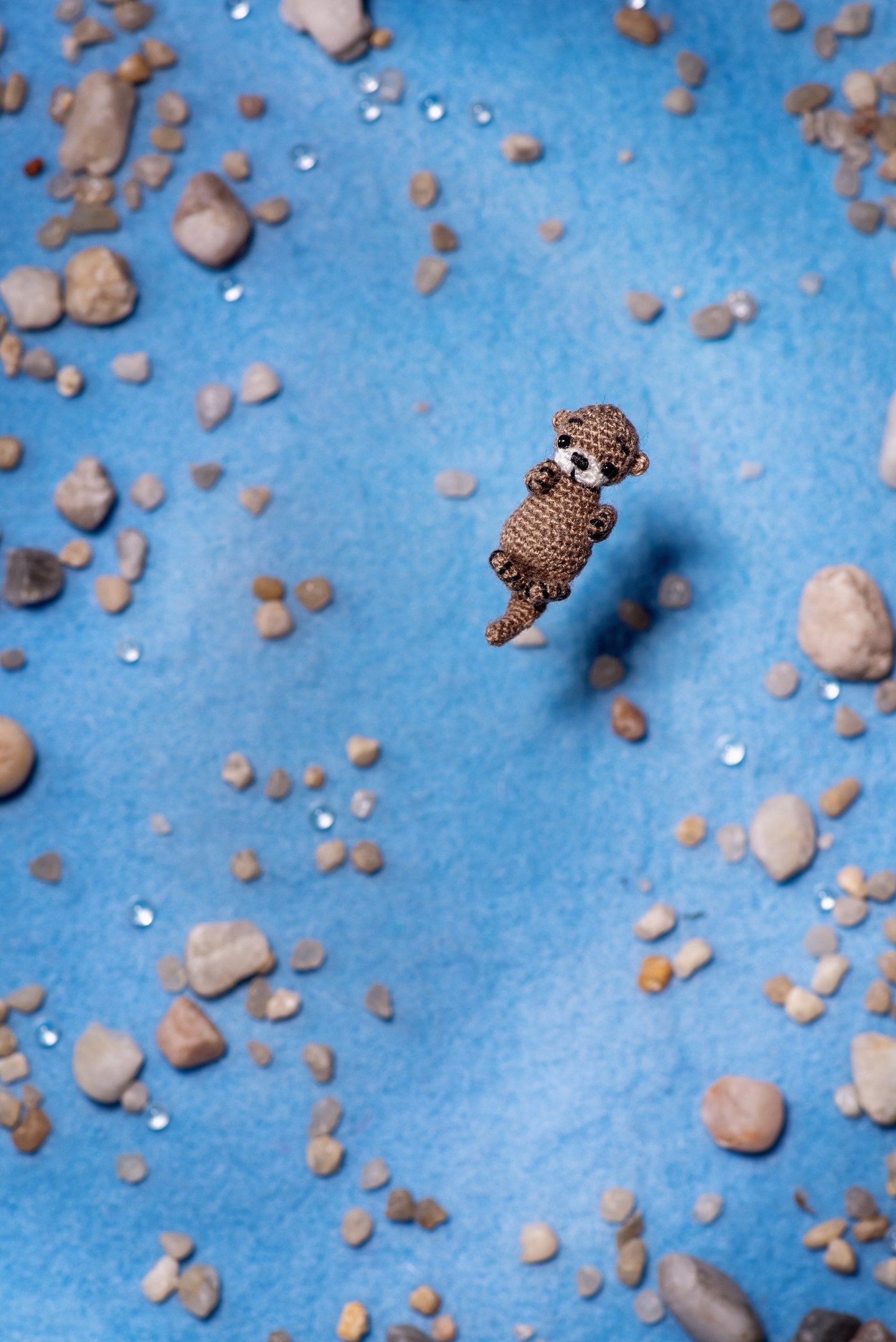 Miniature crochet otter floating on blue with stones and sparkling beads.