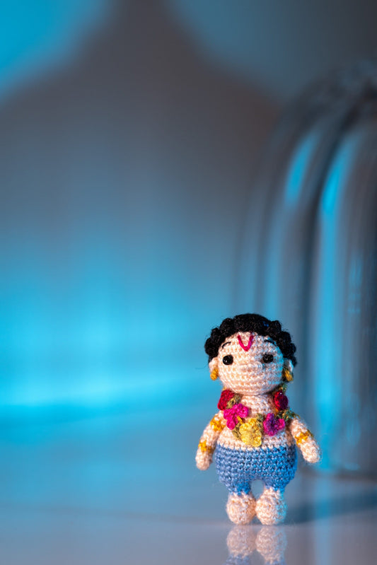 Miniature crochet Lord Balarama with floral garland and blue dhoti on reflective surface.