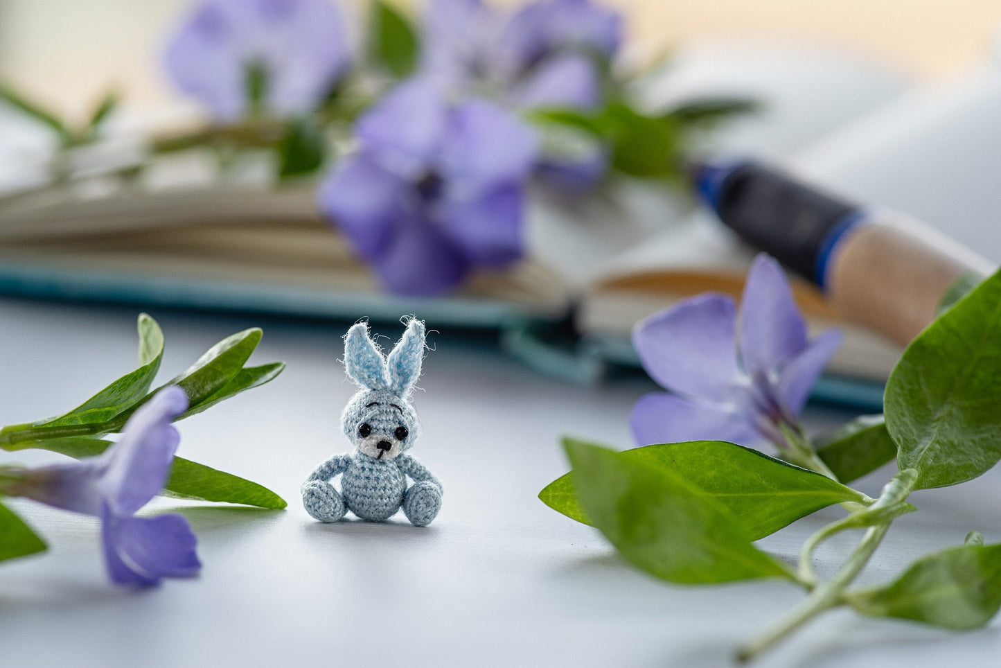 Tiny crochet blue bunny sitting among violet flowers with open notebook and pen
