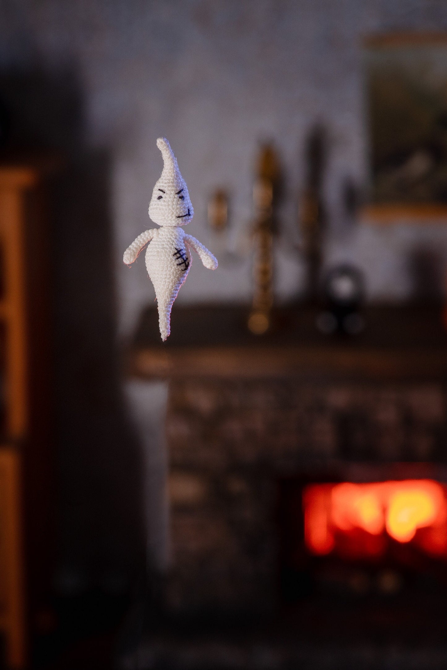 Tiny white crochet ghost floating in front of a warm fireplace in a cozy dollhouse