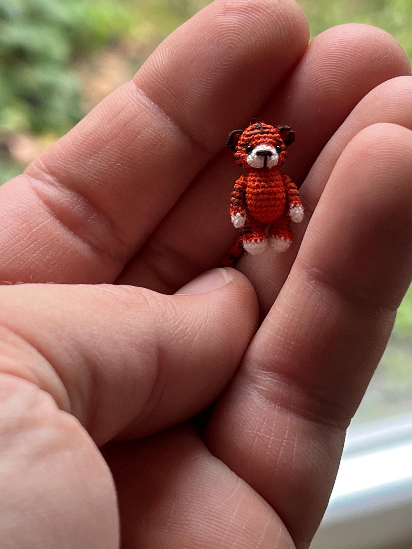 Miniature amigurumi tiger standing on fingertip in nature-inspired scene