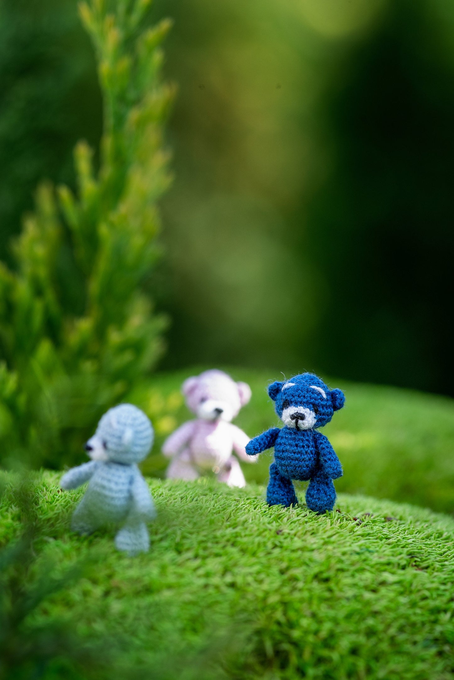 Three knitted teddy bears in blue, white, and gray on a green surface with a blurred natural background.