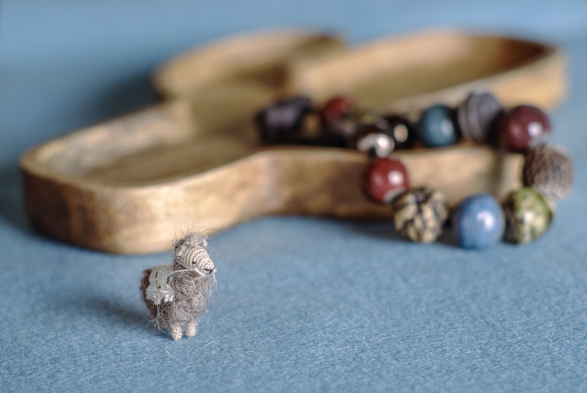 Miniature crochet llama standing on soft blue surface near natural wood tray.