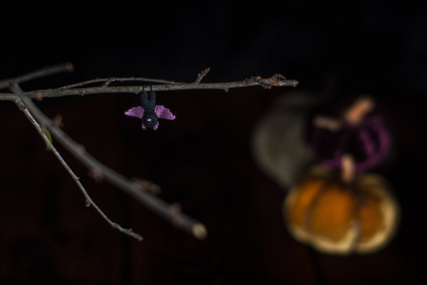 Tiny black and purple crochet bat hanging from a bare twig in moody Halloween lighting.