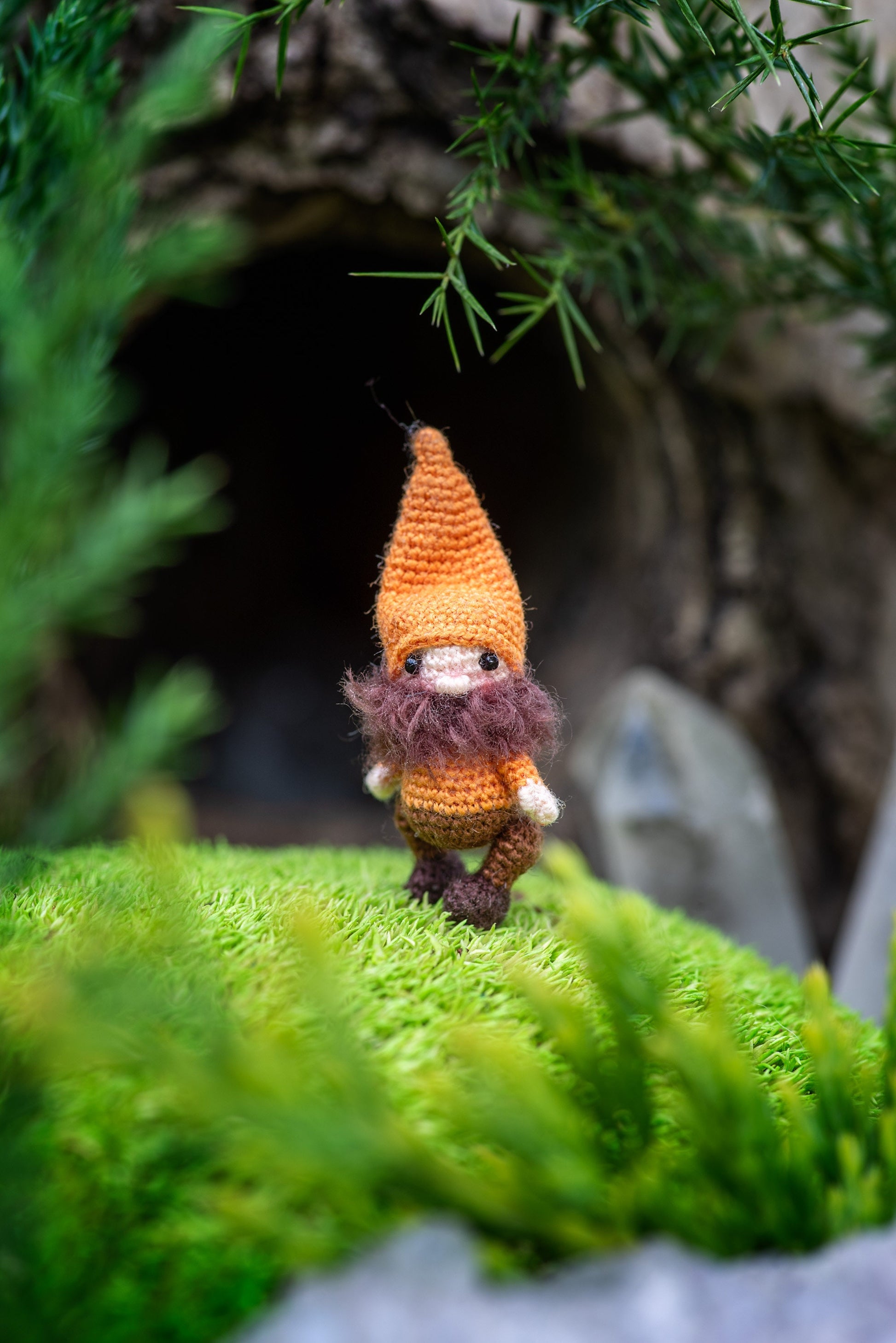 Miniature crochet gnome standing on moss in front of a forest hollow.