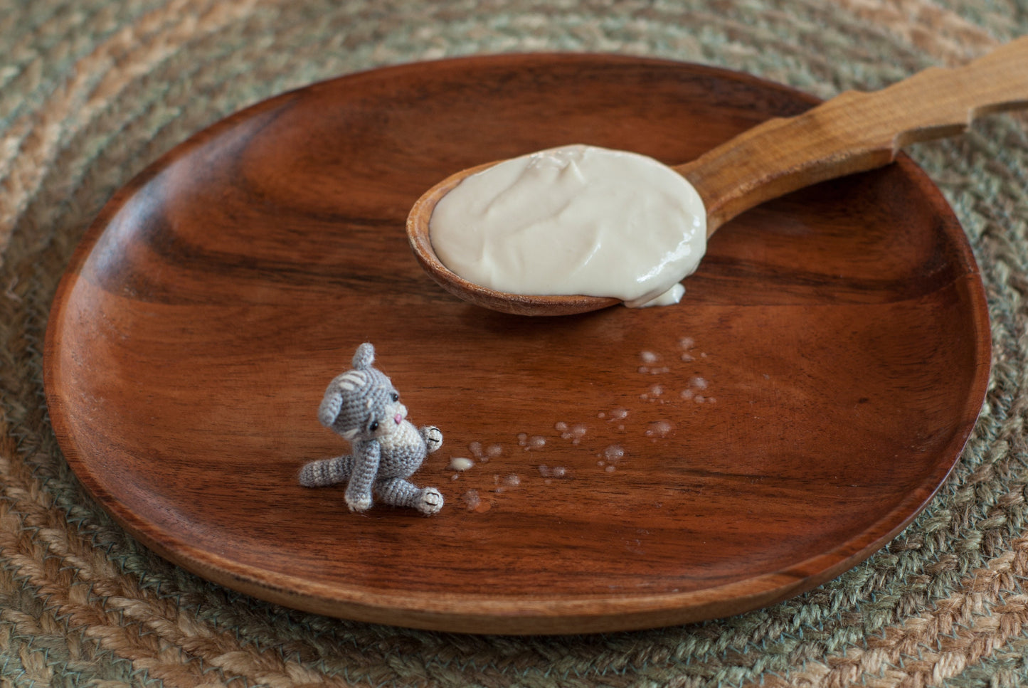 Tiny grey crochet kitten sitting on a wooden plate beside a spoonful of cream.