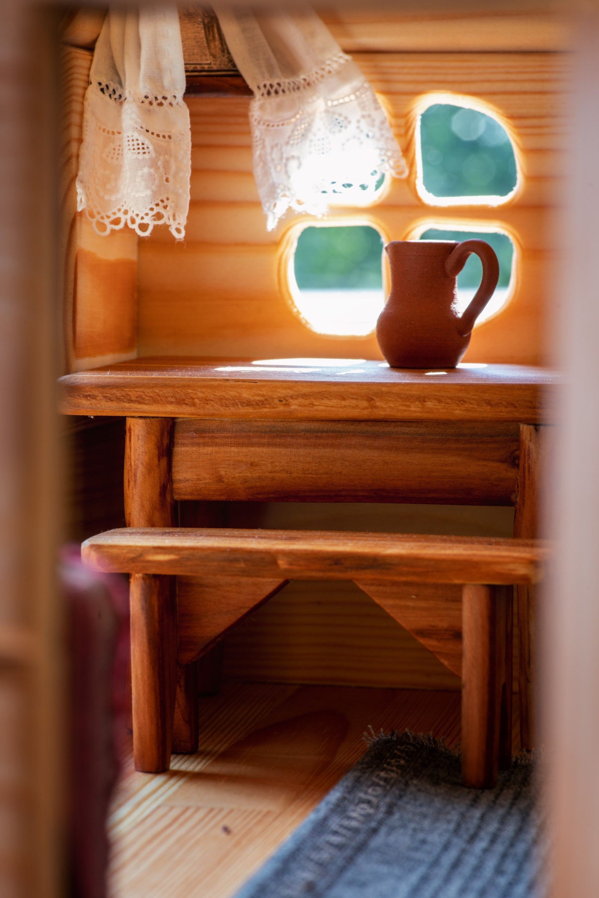 Wooden table with clay jug in rustic dollhouse interior, lace curtains and soft sunlight.