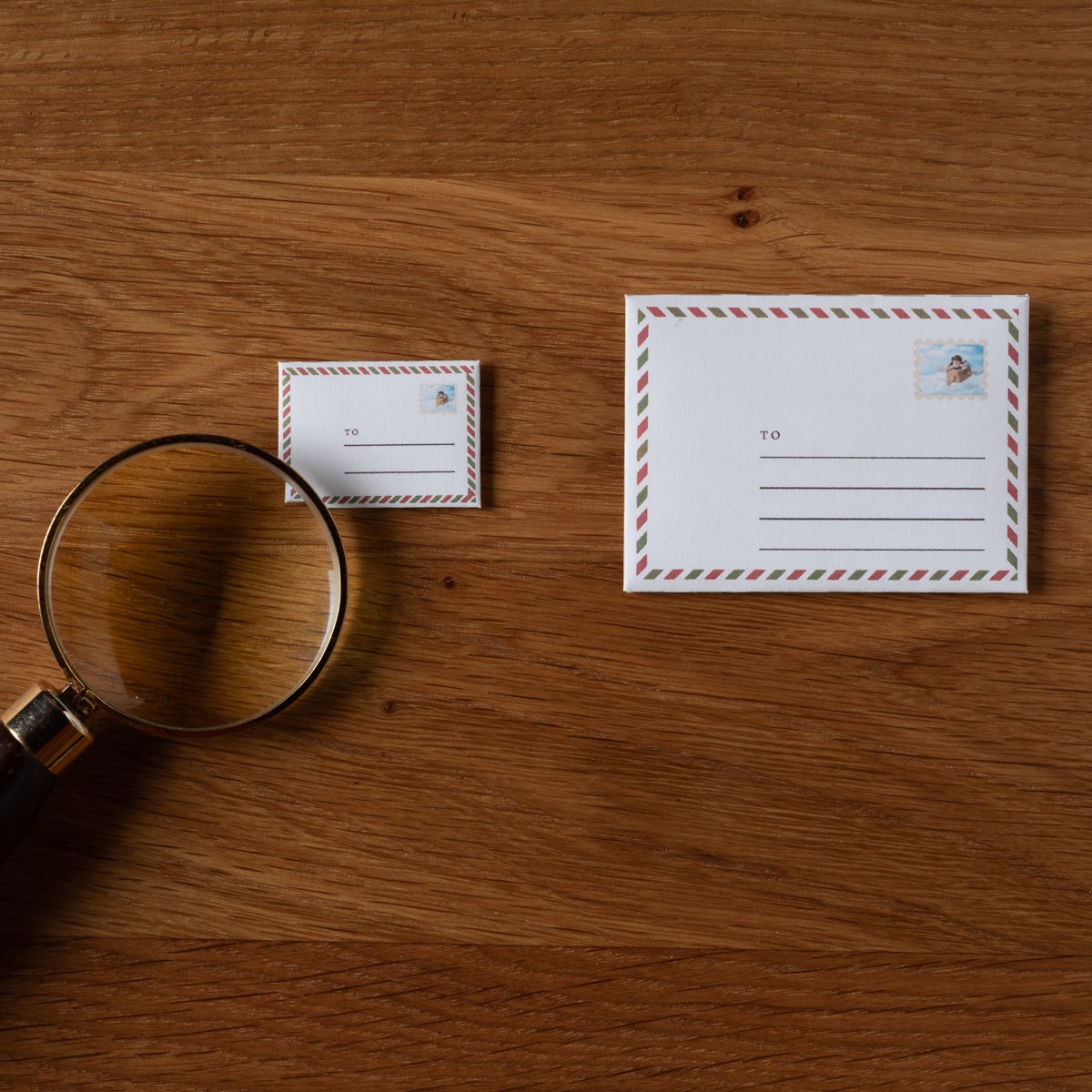 Both sizes on a wooden table — the small envelope and the one twice its size — with a magnifying glass and a miniature postcard beside them.