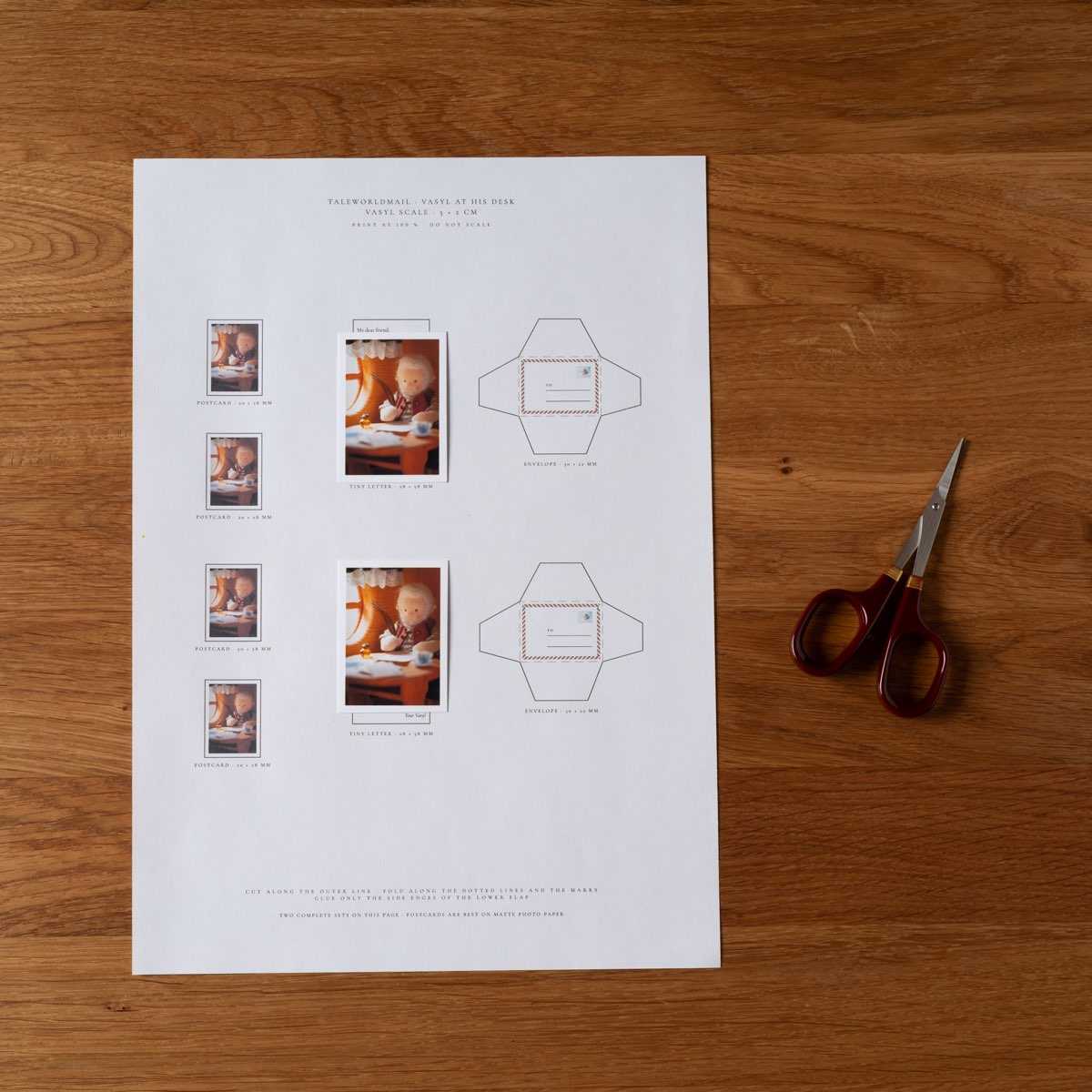 The printed sheet on a wooden table — postcards, tiny letters and two envelope outlines — with a pair of small scissors beside it.