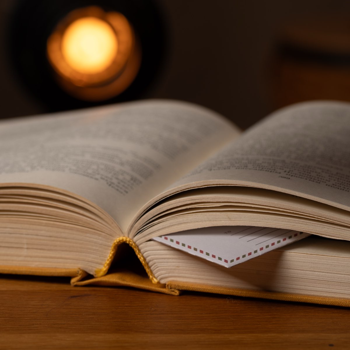 A small airmail-bordered envelope tucked between the pages of an open book, a candle burning behind it.