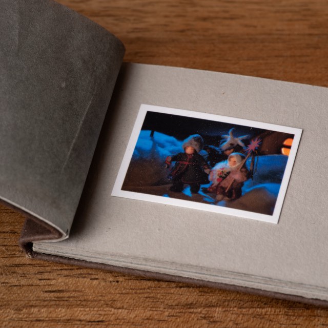 A miniature postcard lying on the grey page of a small open album, its tracing-paper leaf turned back, on a wooden table.