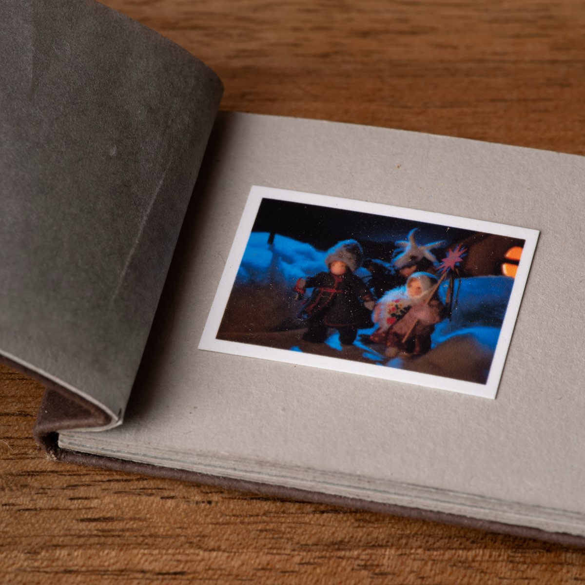 A miniature postcard lying on the grey page of a small open album, its tracing-paper leaf turned back, on a wooden table.