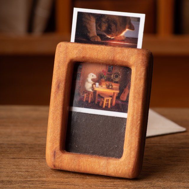 A miniature postcard standing in a small wooden frame, a second card showing above its top edge.