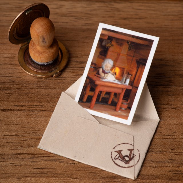 A miniature postcard standing in an open kraft envelope sealed with Vasyl's stamp, the wooden stamp and its brass inkpad lying beside it.
