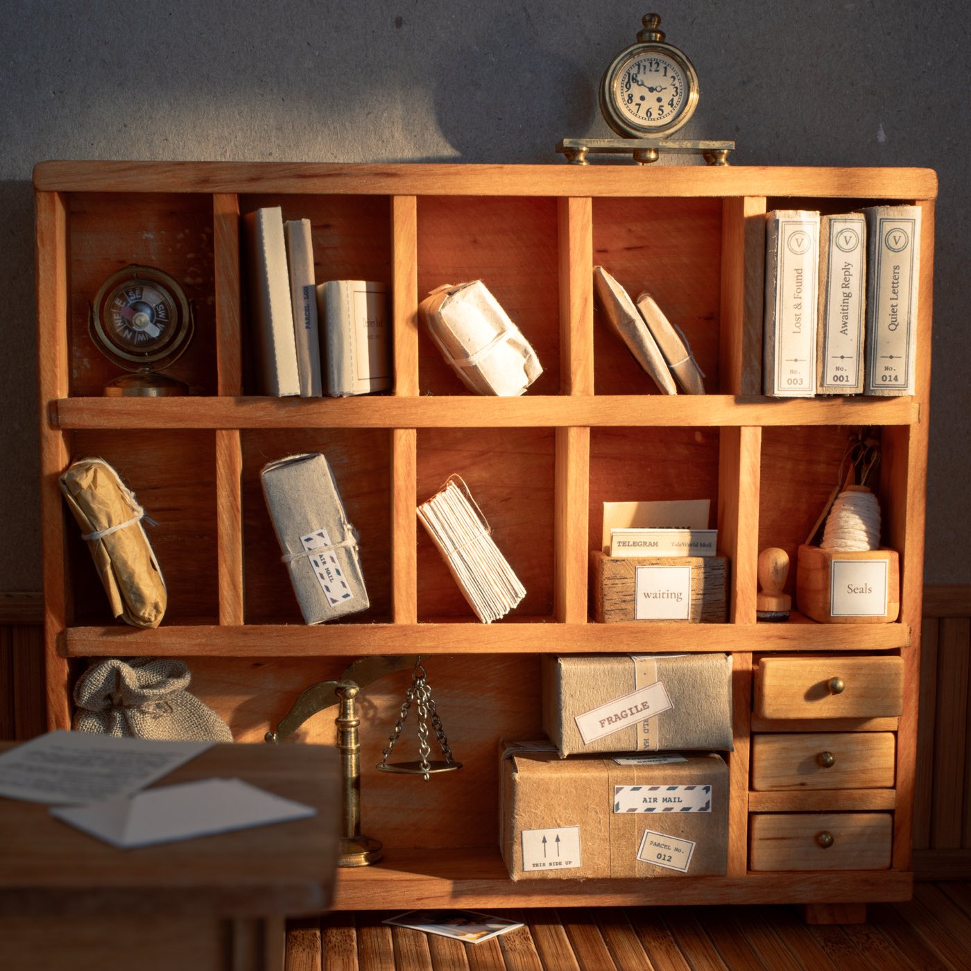 The sorting shelf of a miniature post office: pigeonholes of tied letters and rolled parcels, bundled files marked Lost and Found, Awaiting Reply and Quiet Letters, brass scales, a clock on top, and the corner of a desk with loose envelopes on it.