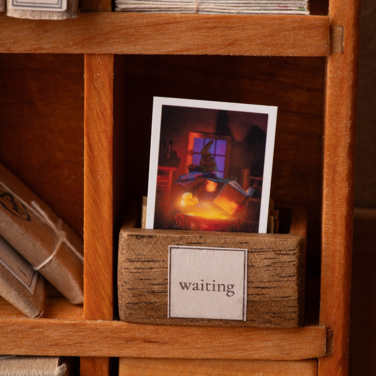 A miniature postcard standing upright in a small wooden box marked «waiting», in one compartment of Vasyl's sorting shelf; tied bundles of letters fill the compartments around it.