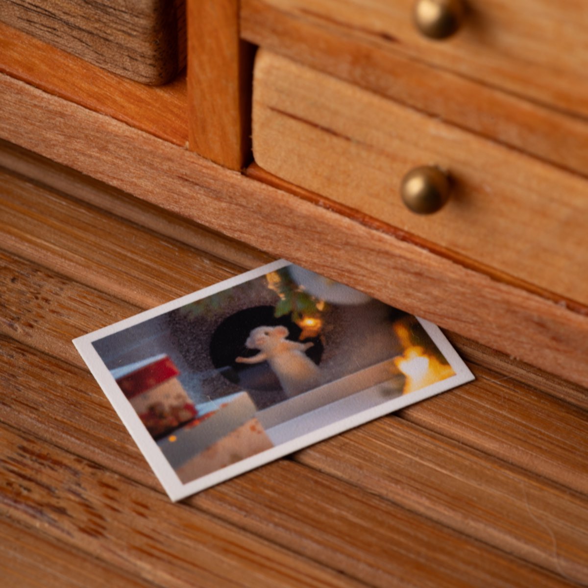 A miniature postcard lying on a plank floor, its upper edge slipped under the bottom of a wooden cabinet with brass knobs.