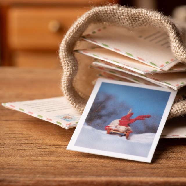 A miniature postcard leaning against a small sack of letters, airmail-bordered envelopes spilling from its mouth onto the table.