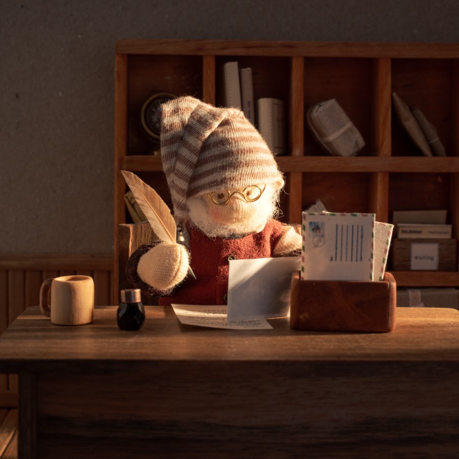 Vasyl, a small gnome in a striped cap, round glasses and a red jumper, sitting at his desk with a letter in front of him and a wooden tray of envelopes at his elbow.